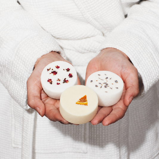 Three handmade aromatherapy shower steamers with rose petals, orange and lavender botanicals, held in cupped hands against a white robe.