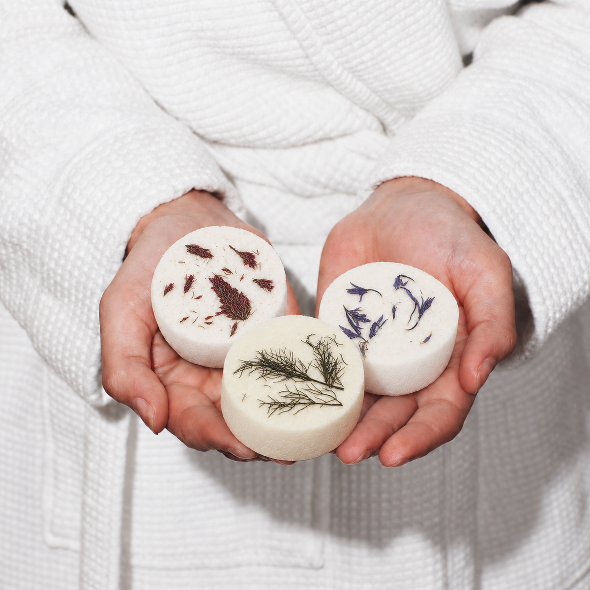 Three Atelier shower steamers, Red Ember, Blue Marine and Emerald Garden, held in cupped hands against a white robe.