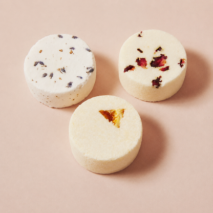 Three handmade aromatherapy shower steamers with lavender, rose petals and dried orange botanicals on a soft beige background.
